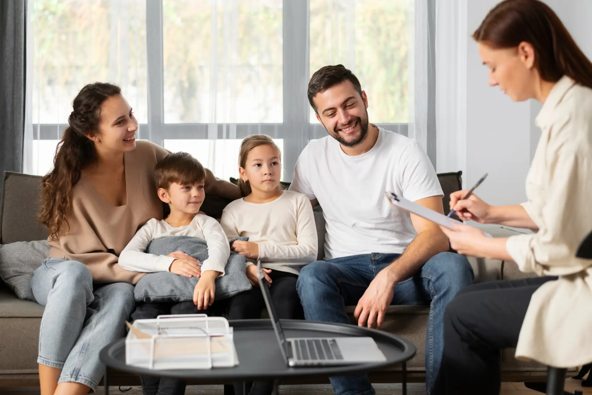 A family of four sits closely together on a sofa while a woman takes notes and observes them in a cozy living room setting